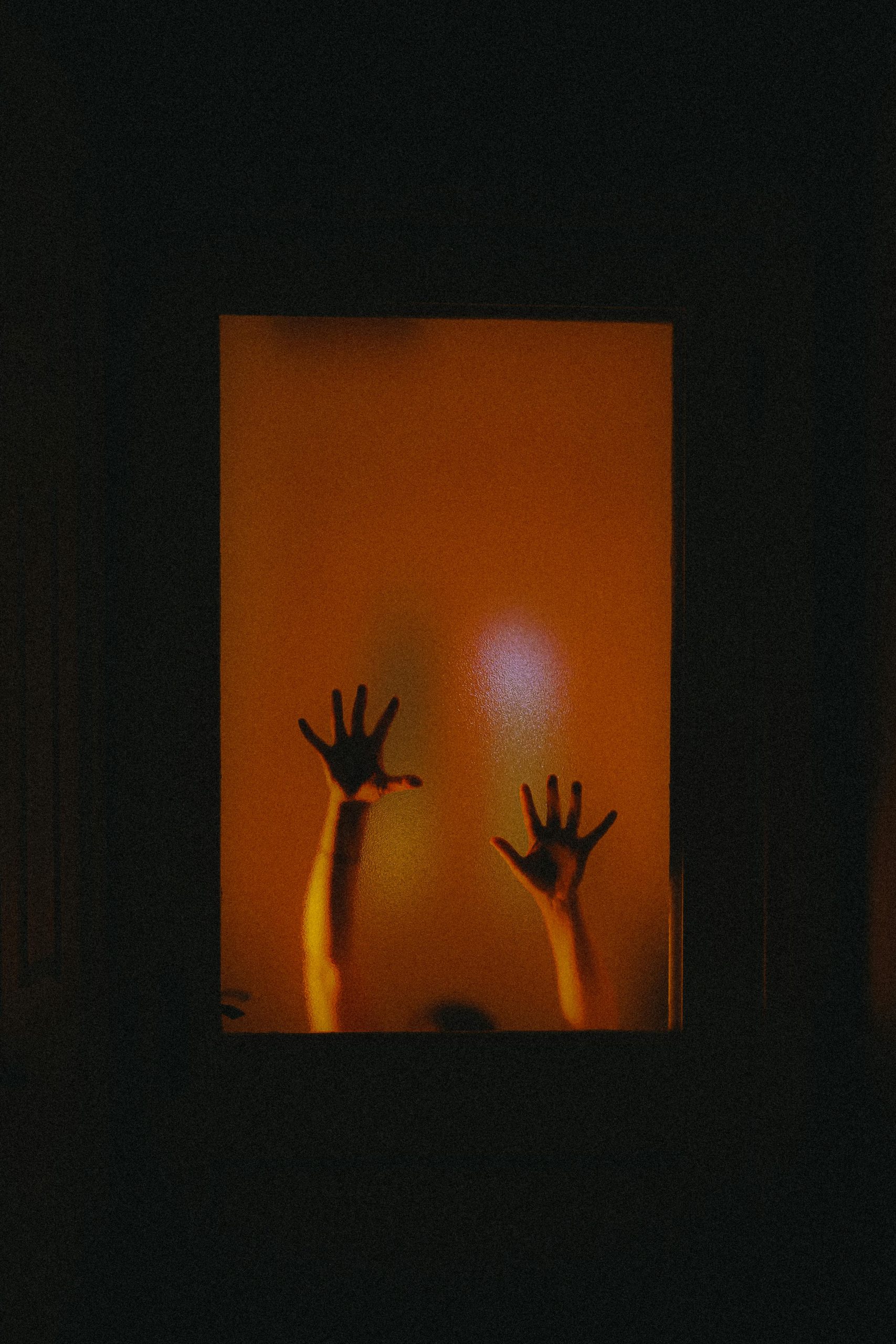 Mysterious silhouette of raised hands behind frosted glass with a warm glow.